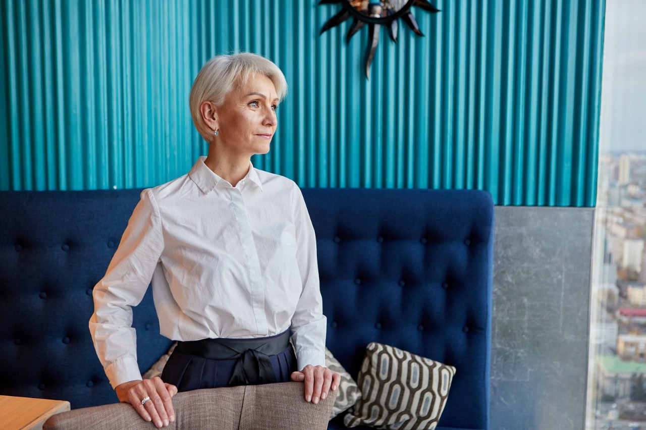 Gray haired lady in elegant clothes standing near couch and touching chair back while looking away dreamily in modern workspace
